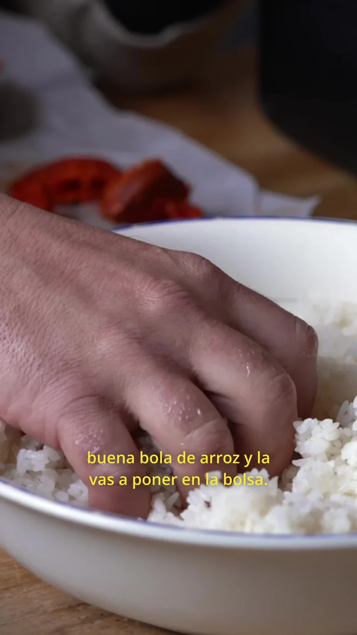 Wet your hand, take a ball of rice, and place it in a plastic bag. Make a small hole, add sobrasada, and close it like a ball. Press firmly and shape it into a triangle using the plastic. Wet your hand to prevent sticking and remove the onigiri.